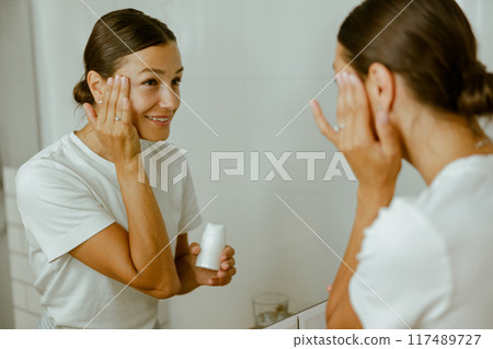 A Woman is Applying Her Skincare Routine in Front of a Mirror, Focusing on SelfCare and Beauty 117489727