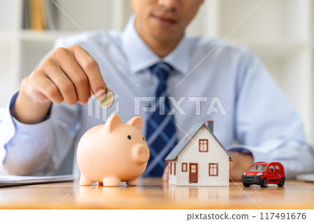 Close up hand of a businessman putting coin in a pink piggy bank, with a small house model on the table, saving or houise mortgage concept 117491676