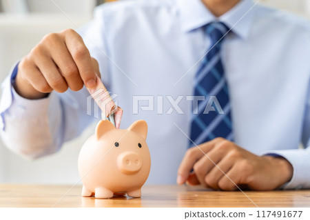 Close up hand of a businessman putting mondy in a pink piggy bank, with a small house model on the table, saving or houise mortgage concept 117491677