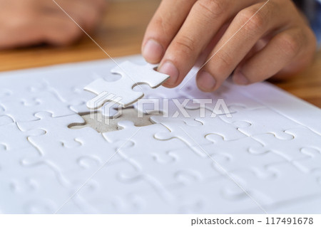 Close up hand of a man holding a white jigsaw puzzle piece trying to put it in an empty slot on the puzzle Close up hand of a man holding a white jigsaw puzzle piece trying to put it in an empty slot on the puzzle 117491678