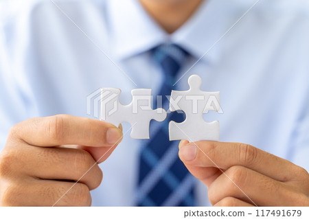 Close up hands of a businessman holding 2 jigsaw puzzle piecess trying to put them together 117491679