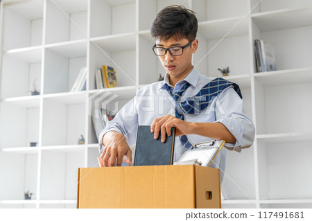 Sad young businessman putting his stuff in a box before leaving his office after his resignation Sad young businessman putting his stuff in a box before leaving his office after his resignation 117491681