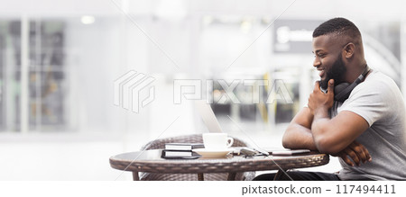A man sits at a table in a cafe, working on his laptop while smiling. He has a cup of coffee and headphones around his neck. 117494411