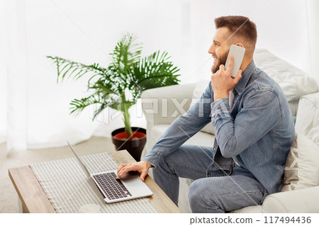 A man is sitting on a couch, engaged in conversation on his cell phone. A man is sitting on a couch, engaged in conversation on his cell phone. 117494436