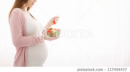 A pregnant woman is standing, holding a bowl of salad. She is looking down at the bowl, likely about to eat. Her baby bump is visible, indicating her pregnancy, cropped, copy space 117494437