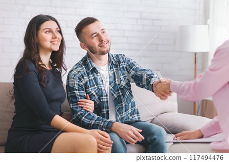 A couple sits on a couch during a therapy session. The man is shaking hands with the therapist, who is off-camera, while the woman sits beside him, watching 117494476