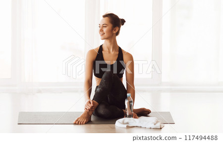 A woman in workout attire sits on a yoga mat outdoors, holding a bottle of water. She appears focused and ready for her exercise routine. A woman in workout attire sits on a yoga mat outdoors, holding a bottle of water. She appears focused and ready for her exercise routine. 117494488