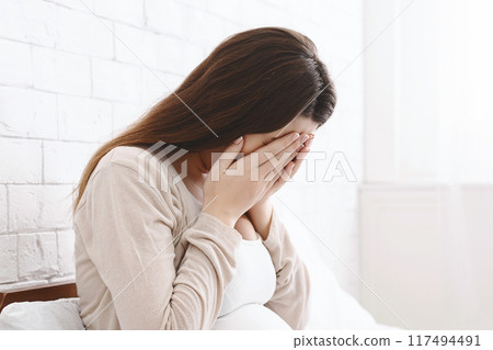 A young woman appears to be distressed as she sits on her bed with her face buried in her hands. The room is bright, with sunlight illuminating the white brick wall behind her. A young woman appears to be distressed as she sits on her bed with her face buried in her hands. The room is bright, with sunlight illuminating the white brick wall behind her. 117494491