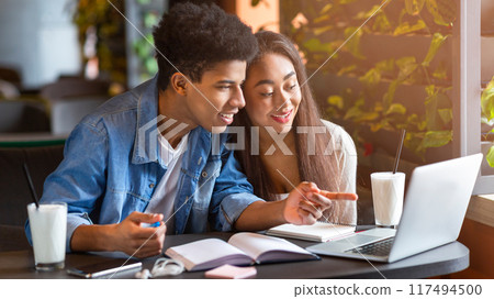 A young loving black couple sits at a table in a bustling cafe, focused on the screen of a laptop. They appear engaged in conversation as they review information on the device. 117494500