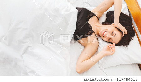 A woman is seen lying down on a bed with a wooden headboard in a simple and cozy room. She appears relaxed and comfortable as she rests on the mattress. 117494513