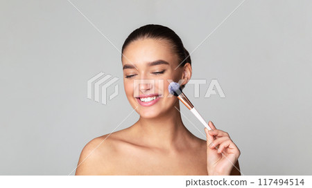 A woman is smiling as she gently brushes her face with a makeup brush, ensuring even application of cosmetics. Her expression denotes contentment and focus during her beauty routine. 117494514