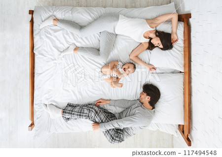 This photo shows a family of three sleeping in a bed together. The parents are on the outside of the bed, while the child is in the middle, top view 117494548