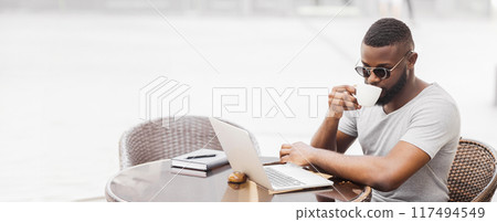 A man wearing sunglasses sits at a cafe table, working on a laptop and drinking coffee. 117494549