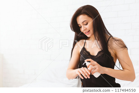 A young woman with long dark hair smiles as she prepares to apply lotion to her hands in a brightly lit, modern bedroom. Dressed in a black tank top, copy space 117494550