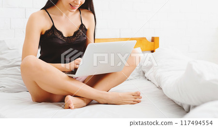 Cropped of woman is seated on a bed, her attention focused on a laptop screen in front of her. She seems engaged in whatever she is viewing on the device. 117494554