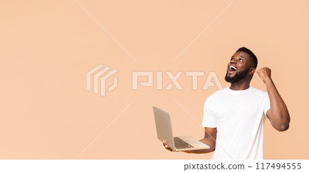 An African American man in a white t-shirt is holding a laptop computer with his left hand and raising his right fist in the air, looking upwards with a joyous expression, copy space 117494555