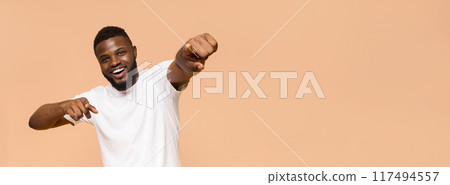 A young Black man with a beard smiles and points with his index finger while wearing a white t-shirt in front of a light peach background. He is looking directly at the camera, copy space 117494557