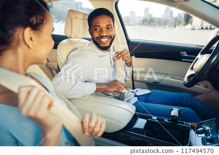 A man in a white shirt and blue jeans is smiling as he fastens his seatbelt in the passenger seat of a car. A woman in a blue shirt is sitting in the front passenger seat and looking towards him. 117494570