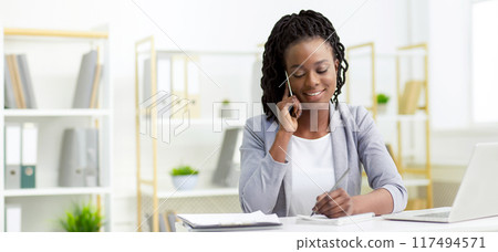 Content young African American lady manager in formal dress, working on a laptop at the office, speaking on the phone. 117494571