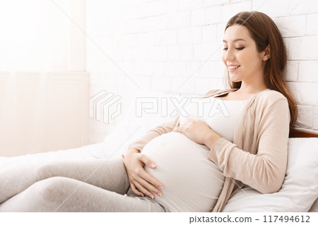 A pregnant woman is seated on a bed, her hand resting on her belly. She appears relaxed and comfortable as she prepares for rest or relaxation, copy space 117494612