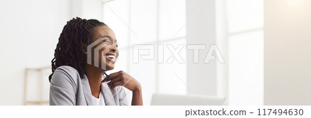 Elated African American professional woman at her desk with a laptop in a modern office space, copy space, web-banner 117494630