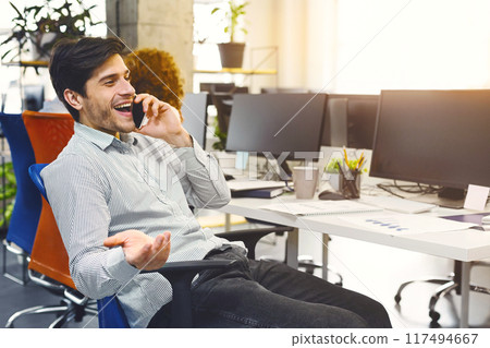 Cheerful man is seated in a chair, engaged in conversation on a cell phone. He appears focused on the call, holding the device to his ear while speaking animatedly. 117494667