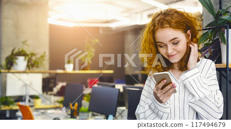 A woman is sitting in an office, focused on her phone screen, reading or typing a message. The office desk is cluttered with papers and a computer monitor is visible in the background. 117494679