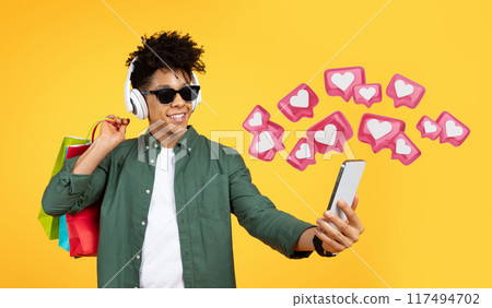 Cool stylish young black guy wearing sunglasses enjoying communication, taking selfie on smartphone, using wireless headphones, listening to music, yellow background. Dating mobile app 117494702
