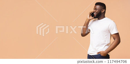 Young Black man talking on his phone while smiling and looking to the left. The man is wearing a white t-shirt and blue jeans and is standing against a beige background, copy space Young Black man talking on his phone while smiling and looking to the left. The man is wearing a white t-shirt and blue jeans and is standing against a beige background, copy space 117494706