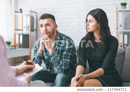 A young couple sits on a couch in an office setting, listening attentively to an off-camera therapist. They appear to be in a serious discussion A young couple sits on a couch in an office setting, listening attentively to an off-camera therapist. They appear to be in a serious discussion 117494711