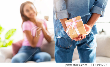 A guy is hiding a gift behind his back, while a girl sits on the couch in the foreground, looking at him. The girl is out of focus, suggesting a surprise. The setting is a casual living room A guy is hiding a gift behind his back, while a girl sits on the couch in the foreground, looking at him. The girl is out of focus, suggesting a surprise. The setting is a casual living room 117494725