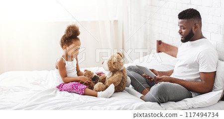 Black father sits on a white bed while his daughter plays with a teddy bear. He is looking down at a tablet in his lap, while she is looking at him. The bed is white and the room is bright. 117494734