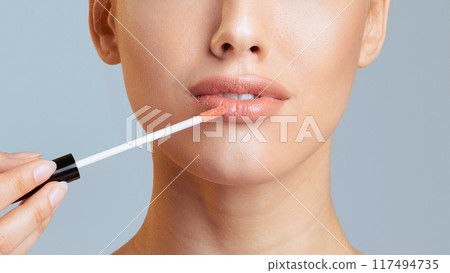 A woman with smooth skin is applying lip gloss to her lips with a clear applicator wand. The background is a light blue, cropped 117494735