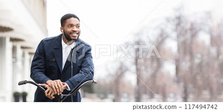 Black businessman is riding his bike to work in a city environment. The atmosphere is urban and energetic, showcasing a practical and eco-friendly mode of transportation. 117494742