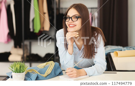 Pretty woman designer is seated at a desk, holding a pen in her hand. She appears focused and engaged in writing or drawing. The scene conveys a sense of concentration and productivity. 117494779
