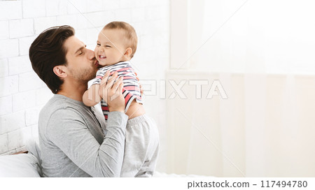 A father is holding his baby daughter, kissing her cheek as she smiles up at him. The father is sitting on a bed with white linens, with a white brick wall visible in the background. 117494780