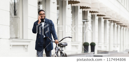An African American businessman is talking on his phone while sitting on a bicycle outdoors. The mood is dynamic and professional, set in an urban environment. An African American businessman is talking on his phone while sitting on a bicycle outdoors. The mood is dynamic and professional, set in an urban environment. 117494806