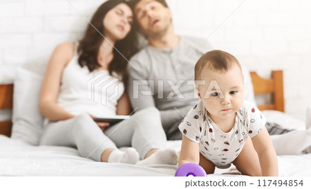 A baby crawls towards its parents who are asleep on a bed. The baby is in focus and the parents are out of focus in the background. A baby crawls towards its parents who are asleep on a bed. The baby is in focus and the parents are out of focus in the background. 117494854