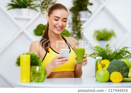 Girl Drinking Detox Juice And Texting On Phone, Sitting In Kitchen Girl Drinking Detox Juice And Texting On Phone, Sitting In Kitchen 117494856