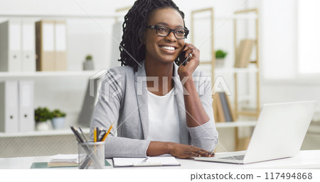 Bright young African American woman in business attire, managing tasks on a laptop at the office, speaking on the phone. 117494868