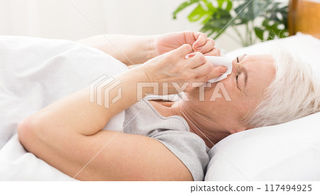 Seasonal flu. Senior woman covering nose with tissue, sneezing in bed, empty space Seasonal flu. Senior woman covering nose with tissue, sneezing in bed, empty space 117494925