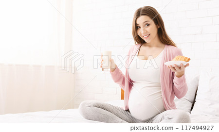 A pregnant woman is sitting comfortably in bed, smiling while holding a glass of milk and a pastry. She appears content and relaxed, showcasing a moment of calm and nourishment during her pregnancy. A pregnant woman is sitting comfortably in bed, smiling while holding a glass of milk and a pastry. She appears content and relaxed, showcasing a moment of calm and nourishment during her pregnancy. 117494927