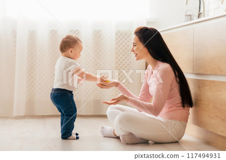 A mother and her toddler are spending quality time together in a bright, modern kitchen. The mother is sitting on the floor, smiling and encouraging her little one who stands in front of her A mother and her toddler are spending quality time together in a bright, modern kitchen. The mother is sitting on the floor, smiling and encouraging her little one who stands in front of her 117494931