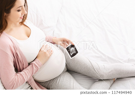 A pregnant woman is seen relaxing on a bed, holding an ultrasound image of her unborn baby. She is comfortably propped against a pillow and gently touching her belly, expressing joy and anticipation. A pregnant woman is seen relaxing on a bed, holding an ultrasound image of her unborn baby. She is comfortably propped against a pillow and gently touching her belly, expressing joy and anticipation. 117494940