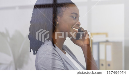Joyful young African American manager in a professional outfit, standing near the office window, engaged in a phone conversation, shot from outdoors Joyful young African American manager in a professional outfit, standing near the office window, engaged in a phone conversation, shot from outdoors 117494954