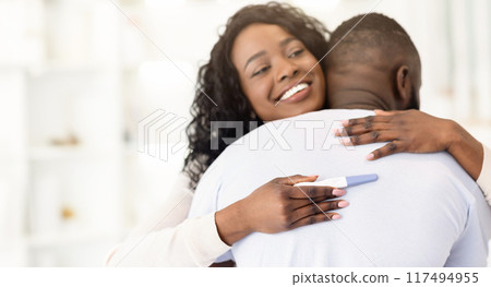 A happy woman embraces her husband from behind, holding a positive pregnancy test, surprising him with the news of their upcoming baby. A happy woman embraces her husband from behind, holding a positive pregnancy test, surprising him with the news of their upcoming baby. 117494955