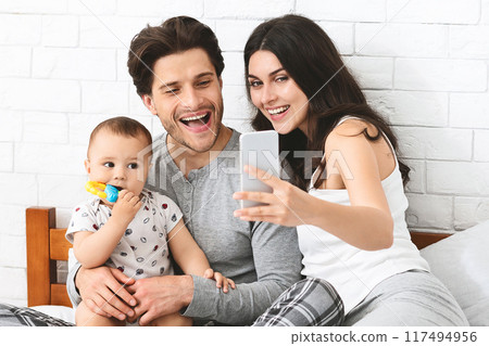 This image shows a happy family taking a selfie in their bedroom. A young man holds his baby son in his lap while his wife takes the picture on a smartphone. 117494956