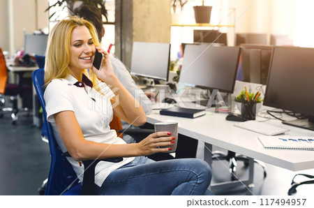 Blonde woman is seated at a desk, engaged in a phone conversation while holding a cell phone to her ear. She appears focused and attentive as she communicates with the person on the other end. Blonde woman is seated at a desk, engaged in a phone conversation while holding a cell phone to her ear. She appears focused and attentive as she communicates with the person on the other end. 117494957