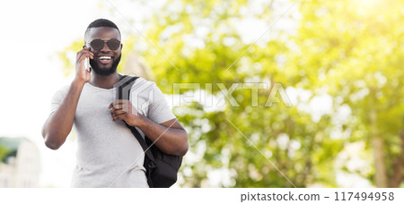 A young man walks through a park while talking on his phone. He is wearing sunglasses and a backpack. A young man walks through a park while talking on his phone. He is wearing sunglasses and a backpack. 117494958