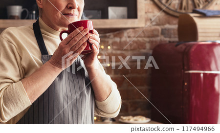 A woman wearing an apron holds a red mug in her hands, smiling and taking a moment to enjoy a warm drink. She is in a cozy kitchen, with brick walls and a red refrigerator in the background 117494966